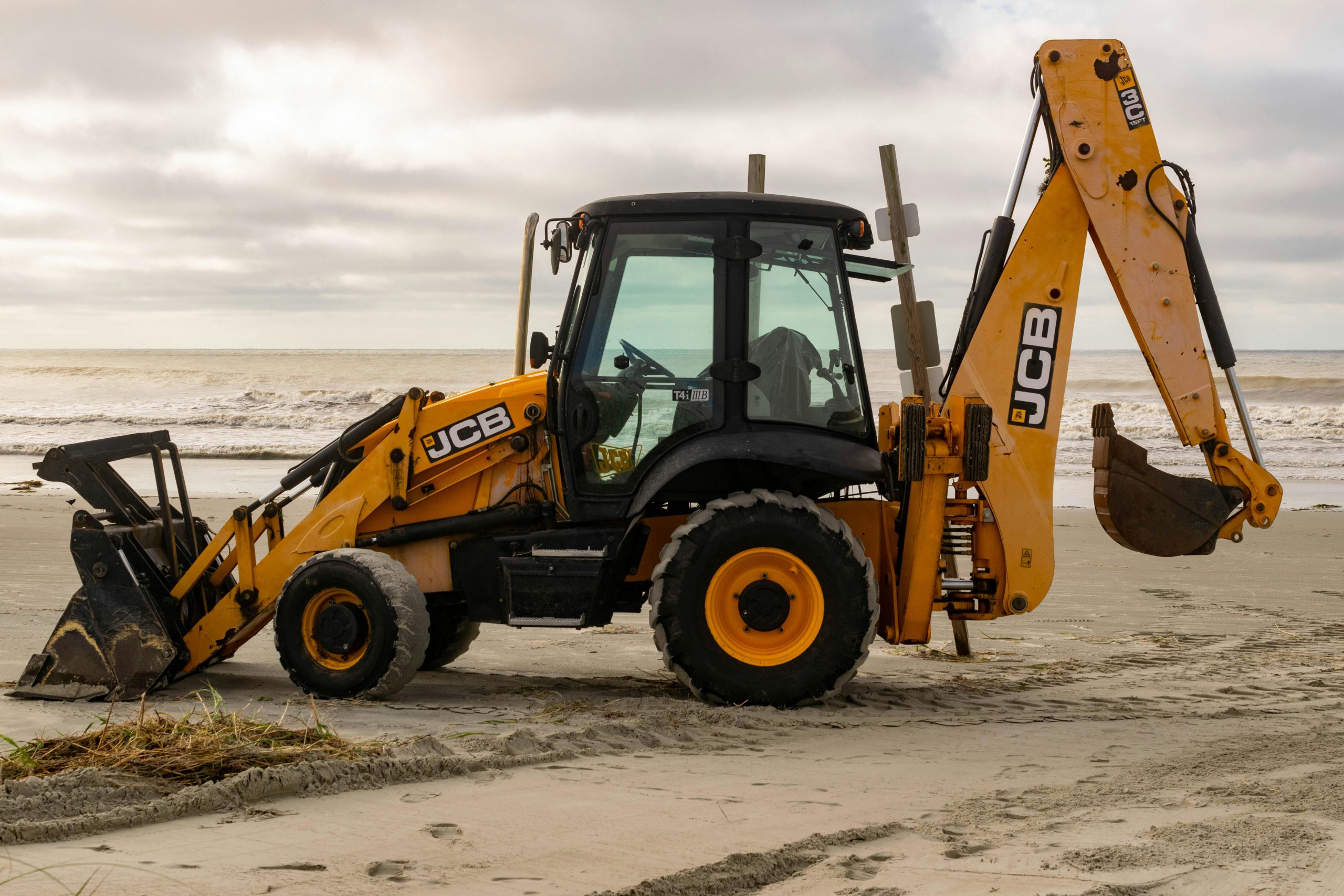 A JCB backhoe parked on the sandy shores of Sunset Beach, North Carolina with ocean waves in the background.
